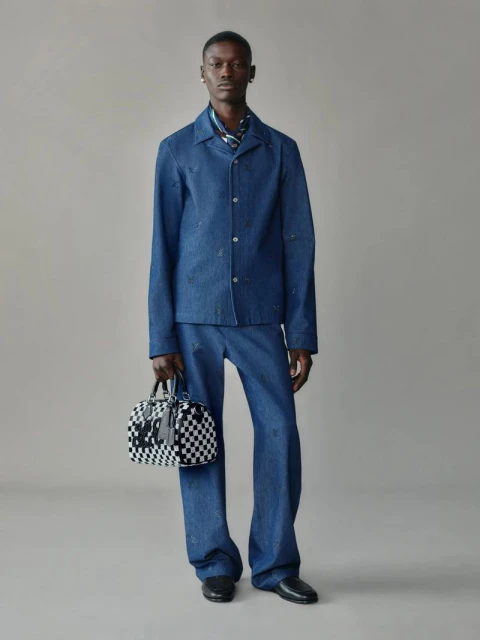 Full-length shot of an adult male fashion model in a denim outfit holding a checkered duffel bag against a plain background.