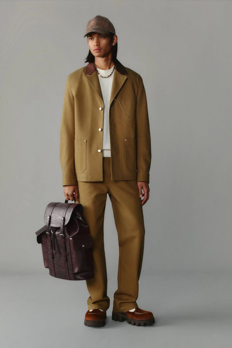 Male model in a tan workwear-inspired jacket and pants, accessorized with a patterned baseball cap and leather backpack in a studio setting.