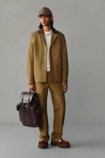 Male model in a tan workwear-inspired jacket and pants, accessorized with a patterned baseball cap and leather backpack in a studio setting.