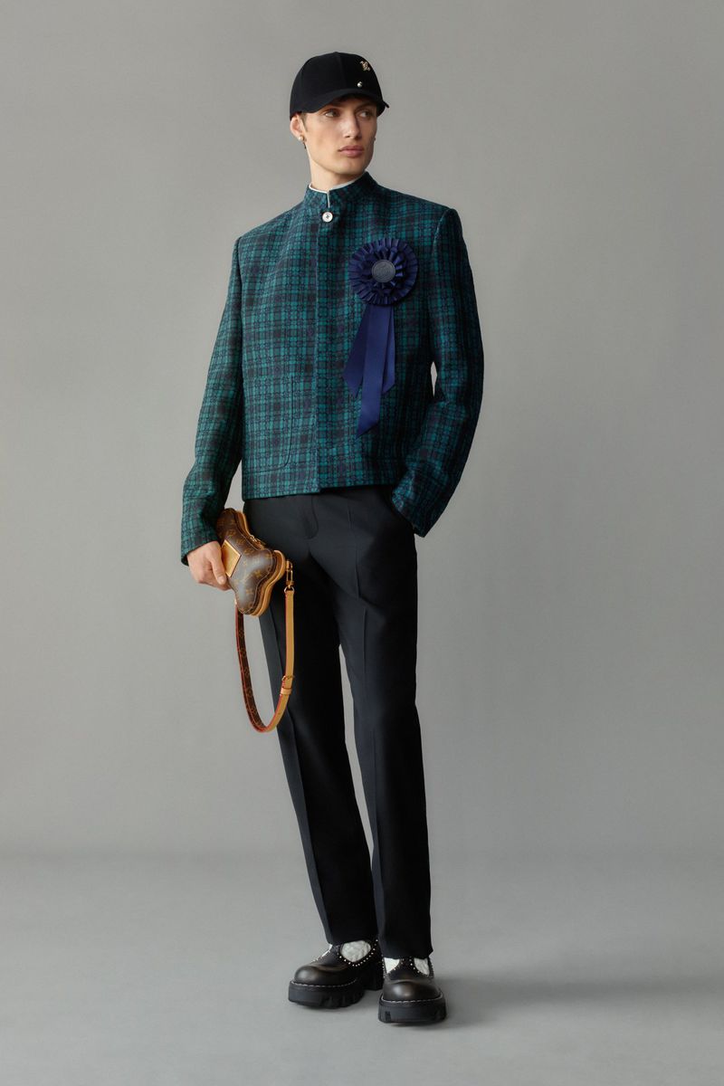 Male fashion model poses in plaid jacket, black pants, cap, and lug-sole shoes, holding a brown handbag in a studio setting.
