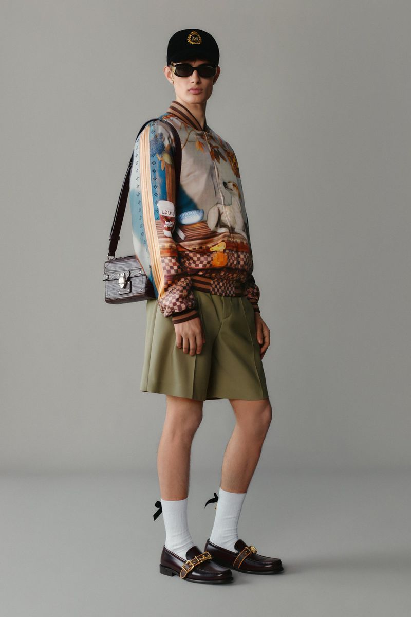 Fashion shot featuring a person wearing a patterned bomber jacket, dark shorts, loafers, a baseball cap, and a shoulder bag.