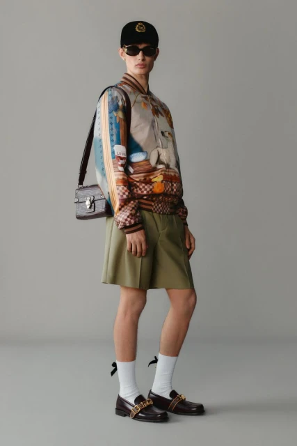 Fashion shot featuring a person wearing a patterned bomber jacket, dark shorts, loafers, a baseball cap, and a shoulder bag.