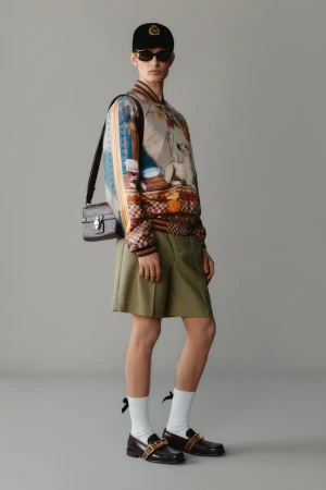 Fashion shot featuring a person wearing a patterned bomber jacket, dark shorts, loafers, a baseball cap, and a shoulder bag.