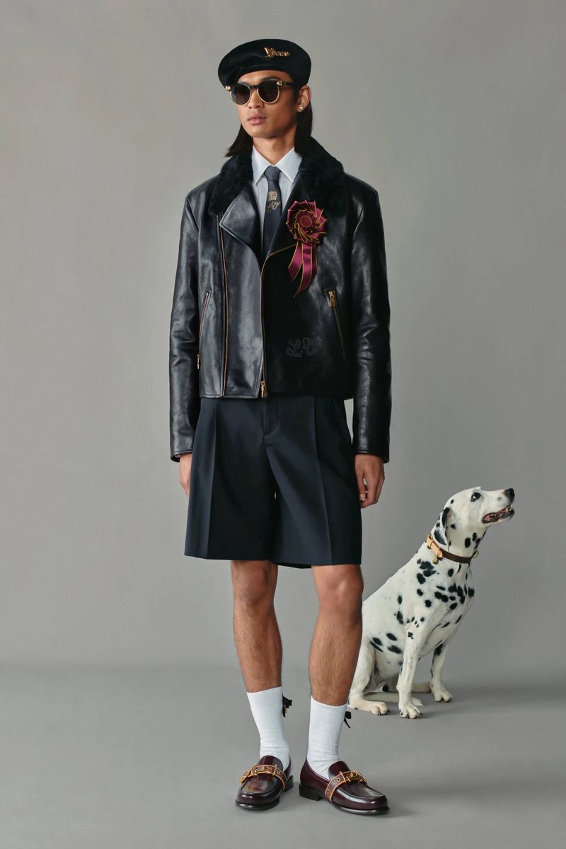 Male fashion model posing with a Dalmatian dog in an indoor studio setting. He wears a leather jacket, shorts, and loafers.