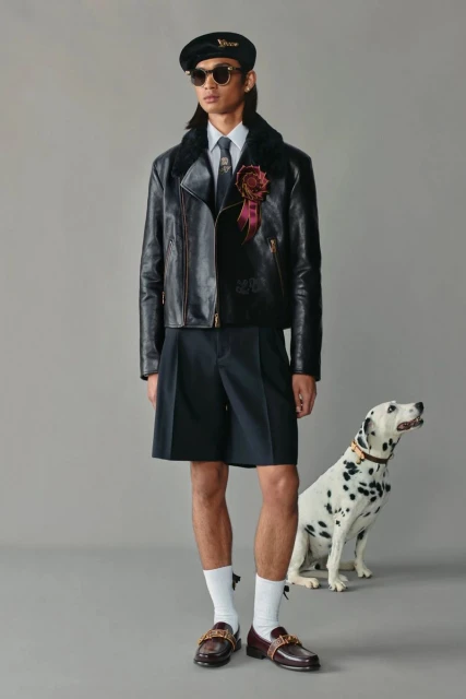 Male fashion model posing with a Dalmatian dog in an indoor studio setting. He wears a leather jacket, shorts, and loafers.