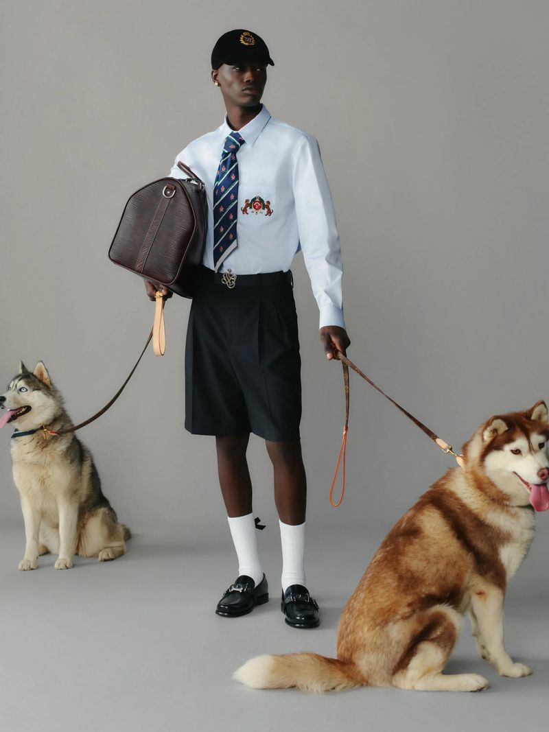 An adult male walks two husky dogs wearing shorts, a button-down shirt, a tie, and a baseball cap, carrying a duffel bag.