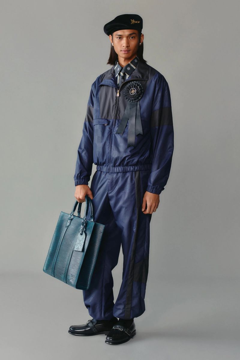 Full-length shot of a slender man wearing a navy tracksuit, a beret, and carrying a blue leather tote bag in a grey studio.