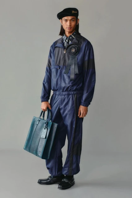 Full-length shot of a slender man wearing a navy tracksuit, a beret, and carrying a blue leather tote bag in a grey studio.