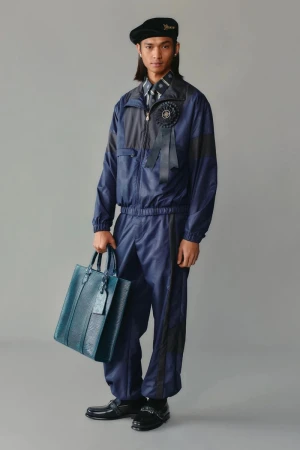 Full-length shot of a slender man wearing a navy tracksuit, a beret, and carrying a blue leather tote bag in a grey studio.