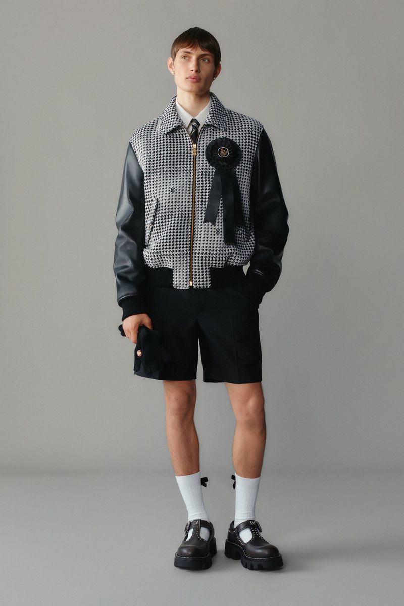 Full-length shot of a male model wearing a black and white jacket with shorts, socks, and Mary Jane shoes in a neutral studio.