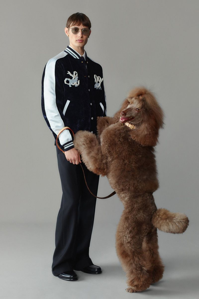 Adult male model poses with a brown standard poodle wearing sunglasses and black and white jacket in a studio setting.