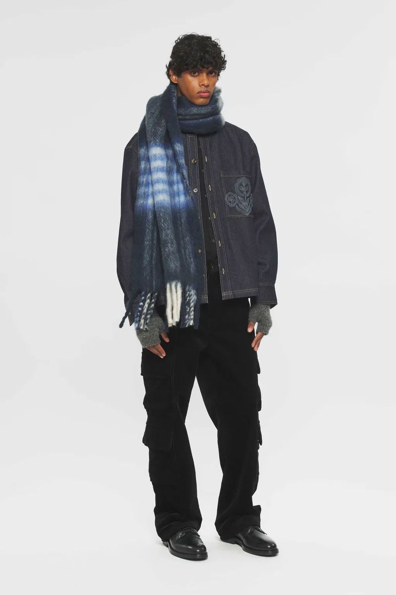 Fashionable adult male in corduroy pants, jacket, and a blue fringed scarf stands against a white background in a studio shot.
