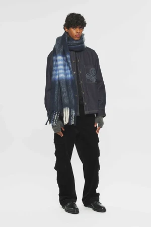 Fashionable adult male in corduroy pants, jacket, and a blue fringed scarf stands against a white background in a studio shot.