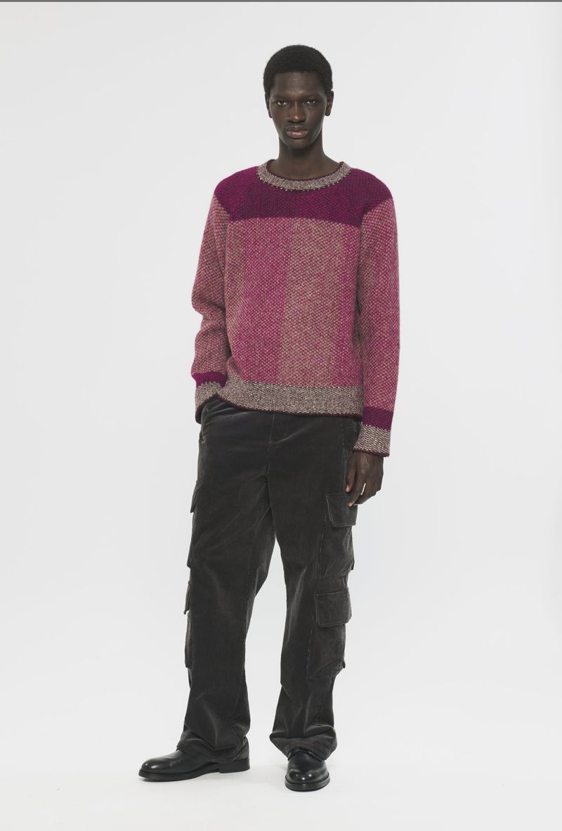 Male model wears a two-tone pink knitted sweater and dark olive corduroy cargo pants against a white backdrop in a full studio shot.