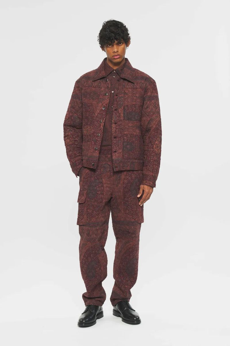 Adult male wearing a patterned red jacket, shirt, and cargo pants poses in front of a white backdrop in a fashion-forward studio shot.