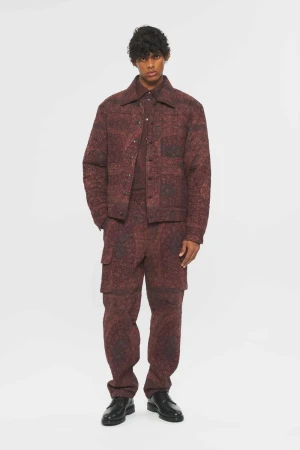 Adult male wearing a patterned red jacket, shirt, and cargo pants poses in front of a white backdrop in a fashion-forward studio shot.