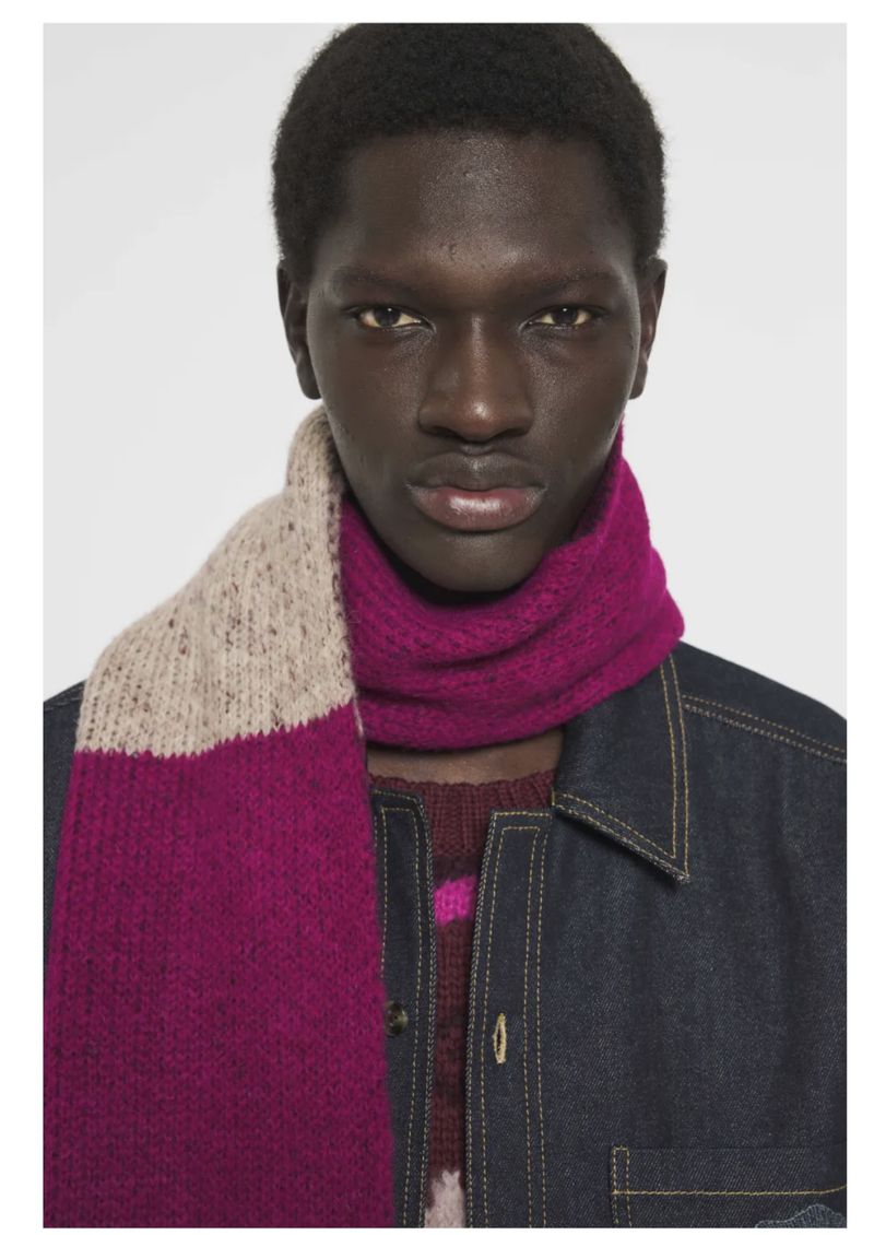 Close-up portrait of Modu Fall wearing a magenta and beige knit scarf and a dark denim jacket against a plain light studio background.