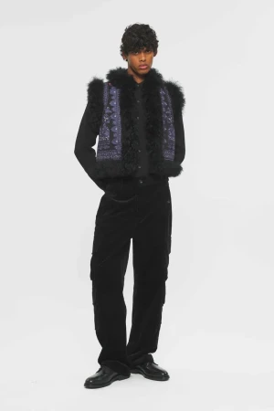 Model Nasir Chaudhary wearing a black fur-trimmed printed vest, black shirt, and trousers against a white seamless background.