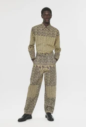 Modu Fall wears a matching patterned beige button-down shirt and trousers with black leather shoes against a white seamless background.
