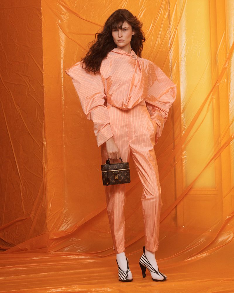 A fashion model in a striped blouse and pants poses in front of an orange backdrop with a Louis Vuitton handbag and striped high heels.
