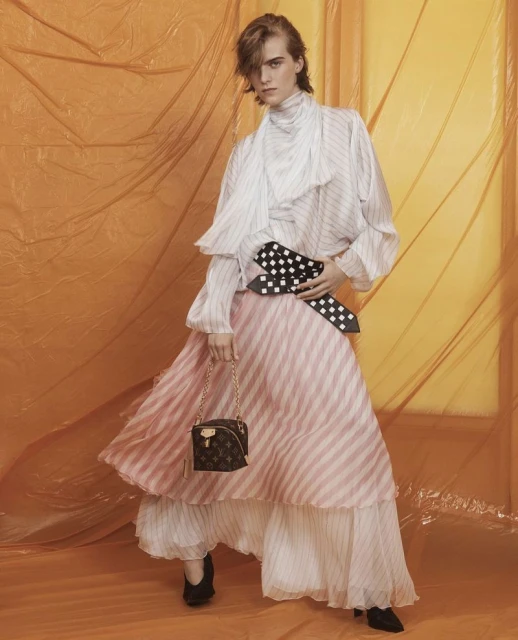 A fair-skinned young adult woman poses in a flowing white and pink striped dress with a Louis Vuitton handbag against an orange background.