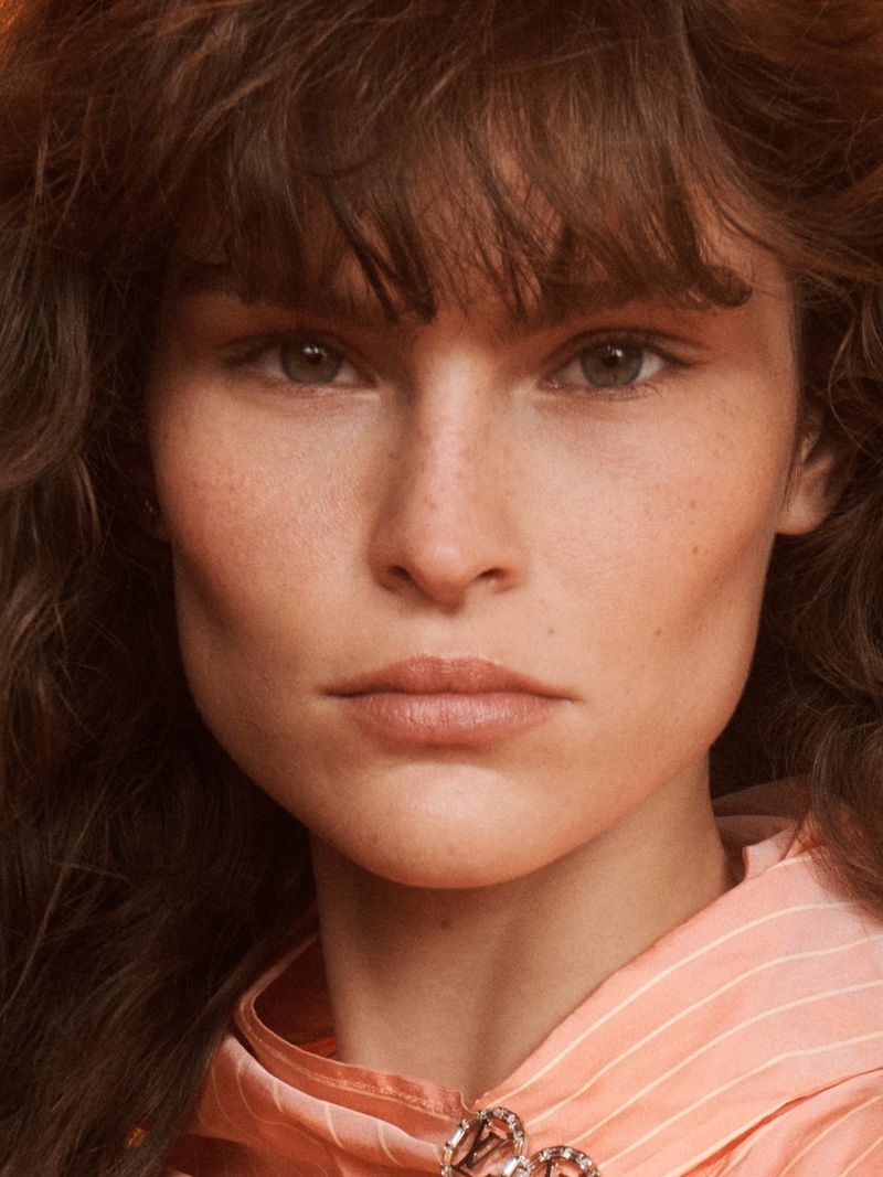 Close-up portrait of a woman with brown curly hair and fair skin, wearing a peach and white collared shirt, shot in soft lighting.