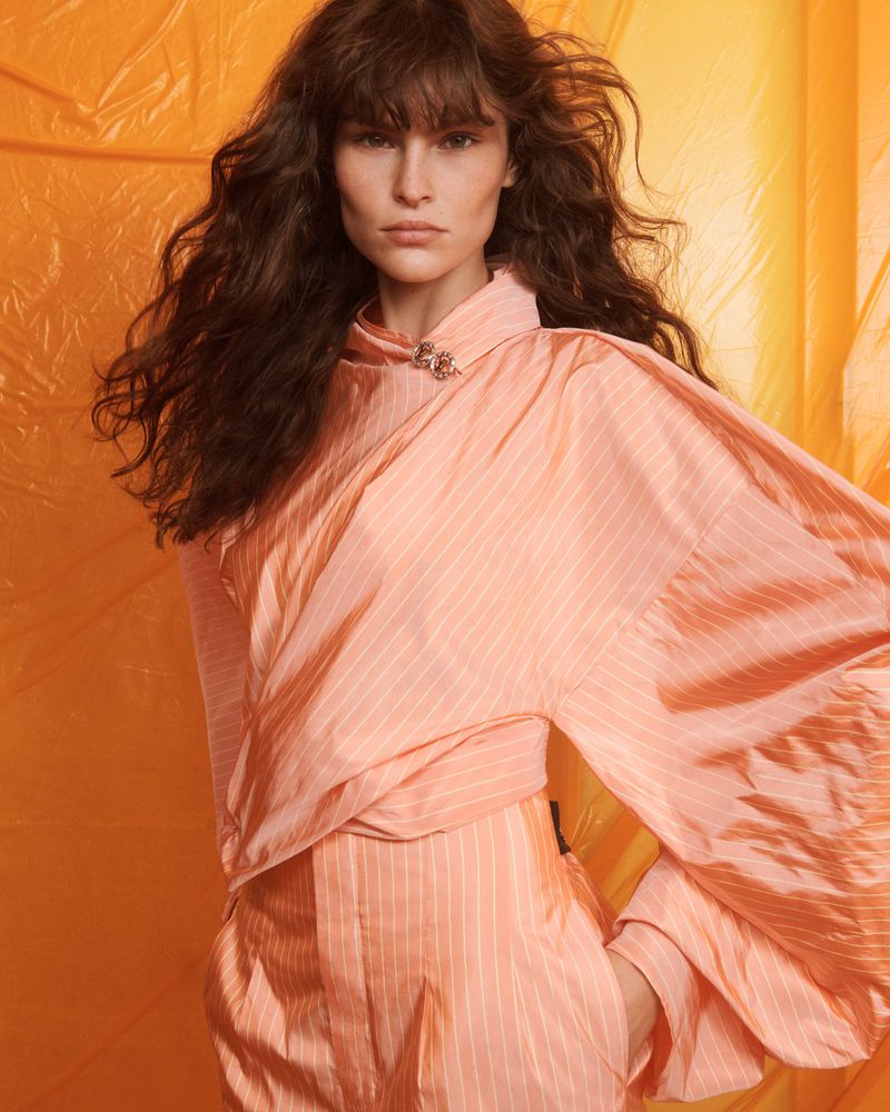 A fair-skinned woman with long brown hair poses in a peach-colored striped outfit against a bright yellow background for a fashion editorial.