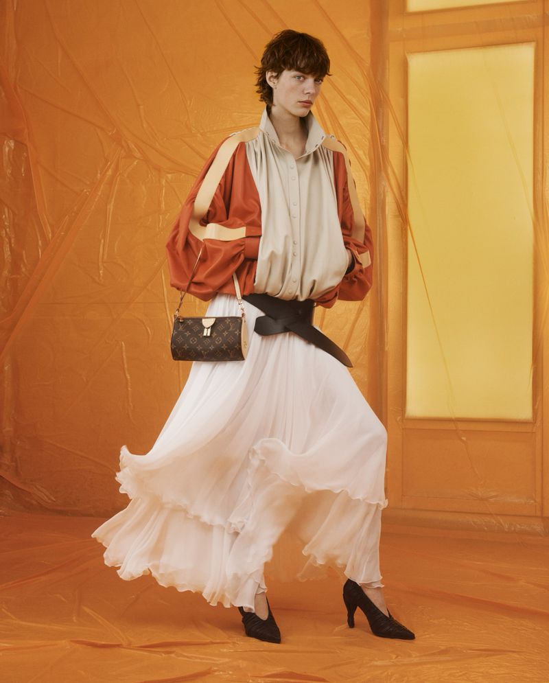 A person in a beige and orange jacket and flowing white skirt poses with a purse in an orange-toned indoor fashion shot.