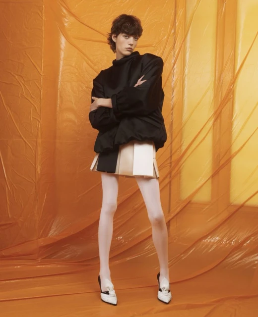 Fashion photography of a model wearing a black jacket, multi-panel mini skirt, white tights, and black and white heels against an orange backdrop.