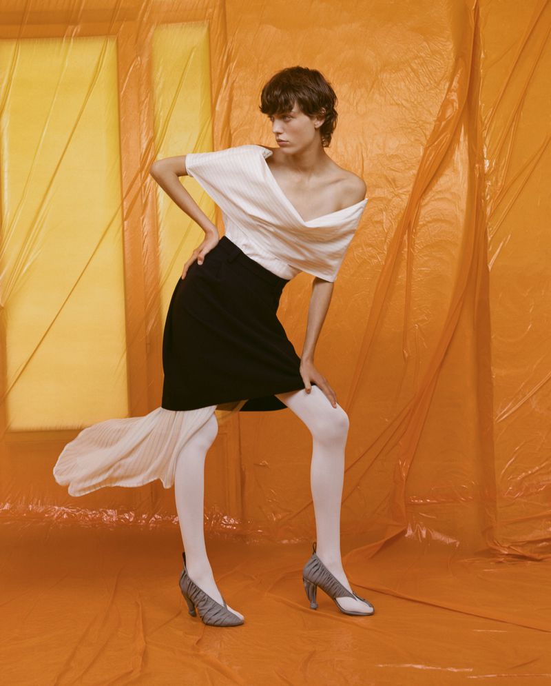 A woman poses in a studio wearing a white blouse, black skirt, and white tights against an orange background with natural lighting.
