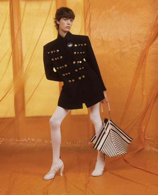 Fashion model poses in a black jacket with gold buttons and white tights, holding a black and white bag against an orange background.