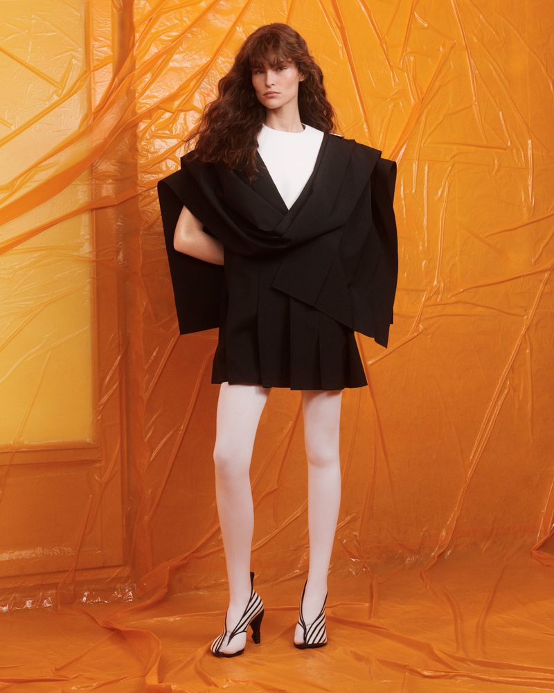 A slender woman poses in a white t-shirt, black jacket, black pleated miniskirt, and striped heels against an orange backdrop.