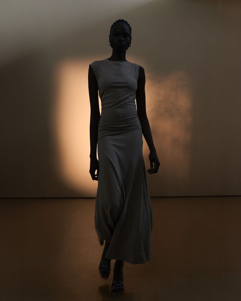 Dark-skinned model in a grey dress walks toward the camera in a dimly lit studio with a soft orange glow highlighting the background.
