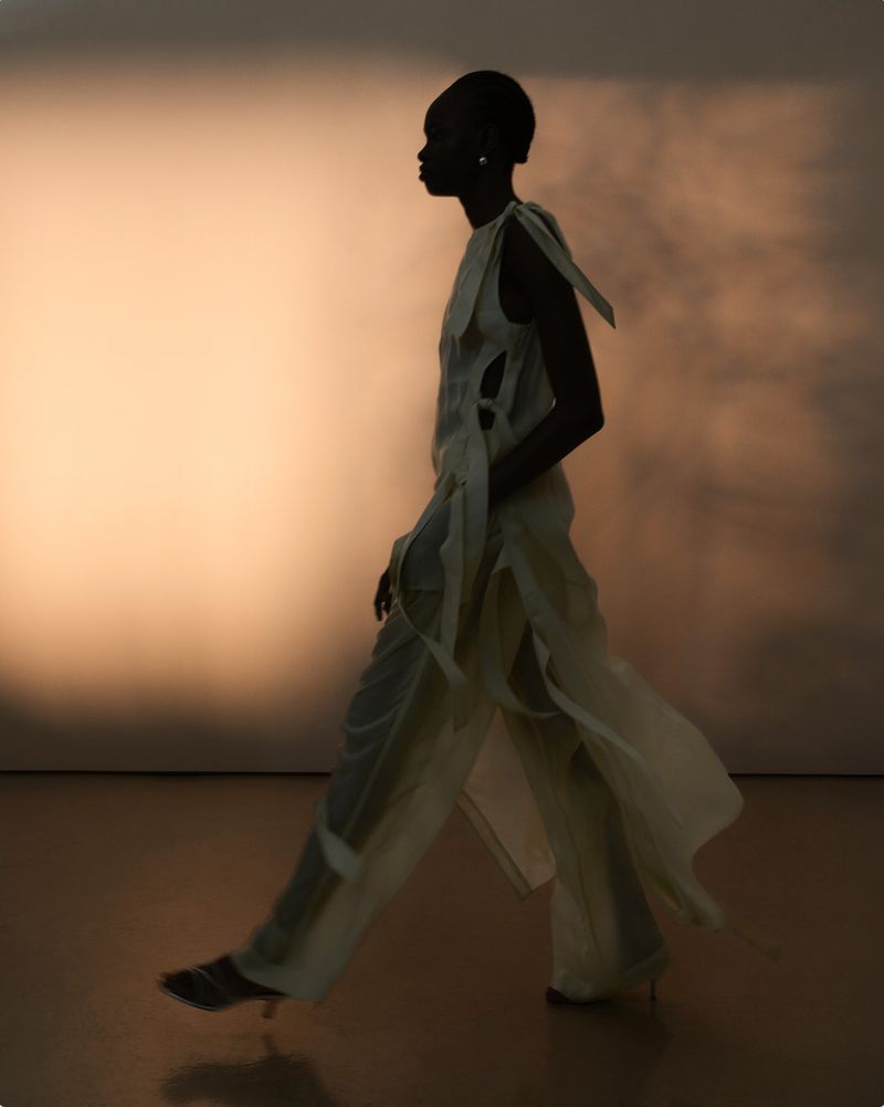 A slender African woman strides confidently in a flowing, light-colored outfit against a soft peach backdrop, radiating elegance and motion.
