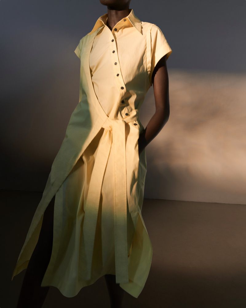 Fashionable woman in yellow button-up dress with short sleeves and pleated skirt posing indoors with hands in pockets.