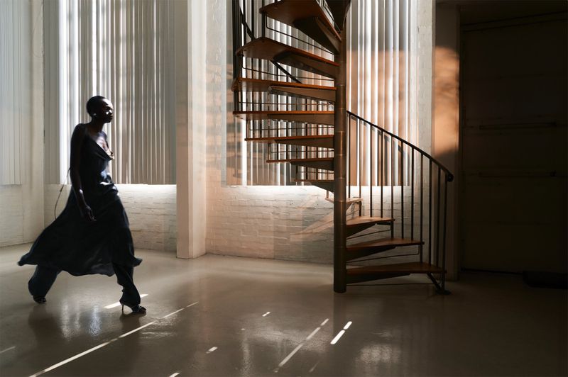Adult female in black dress poses by spiral staircase in interior with natural light shining through blinds, creating bright stripes.