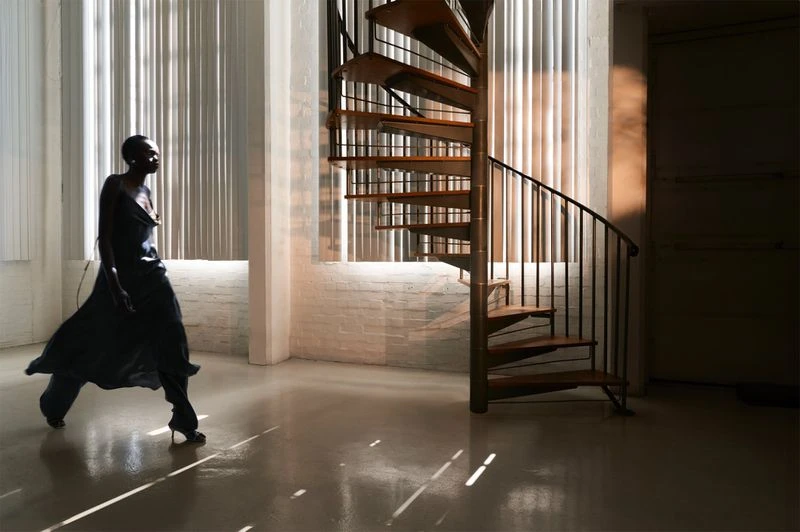 Adult female in black dress poses by spiral staircase in interior with natural light shining through blinds, creating bright stripes.