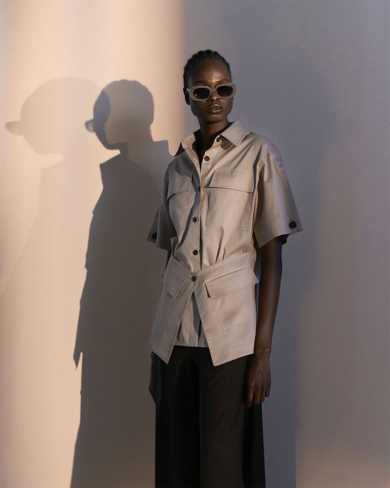 A dark-skinned woman poses in a studio wearing sunglasses, a button-down shirt, and wide-leg pants. A shadow falls behind her.