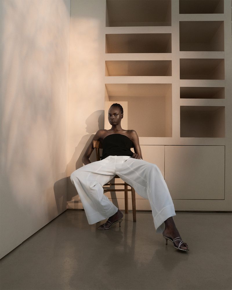 A dark-skinned woman in a black top and white pants sits on a wooden chair against a neutral-toned wall with built-in shelves.