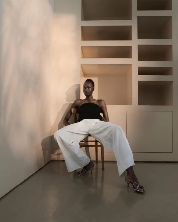 A dark-skinned woman in a black top and white pants sits on a wooden chair against a neutral-toned wall with built-in shelves.