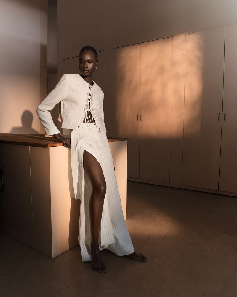 Adult woman in white striped jacket and slit skirt leans against a beige counter in a warm-toned room with light-colored walls and floors.
