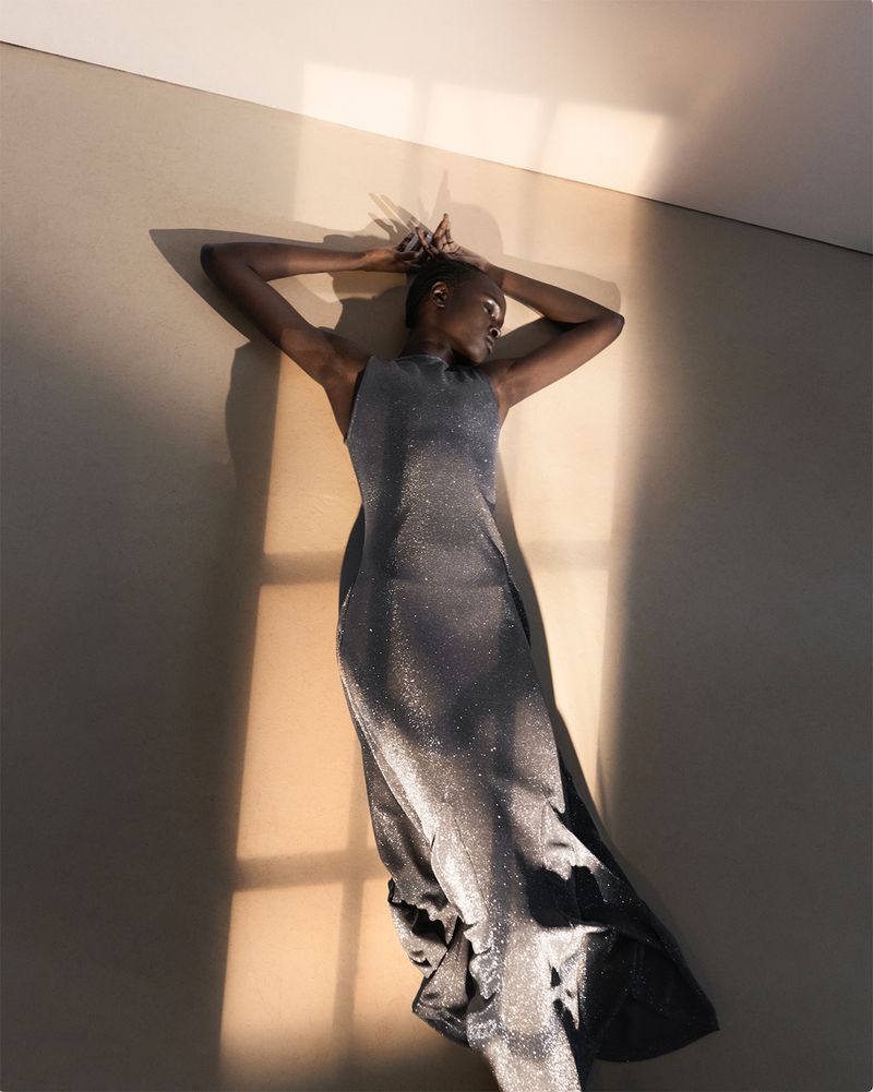 Top view of an adult Black woman lying on her back indoors, with hands above her head, wearing a sleeveless dress with shiny detail.