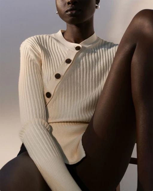 An African woman with a neutral expression poses indoors wearing a cream-colored ribbed sweater with an asymmetrical button design and black tights.