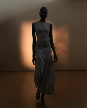 Model Bakhita Lual in a long fitted dress standing against a neutral wall with warm backlighting for Eudon Choi SS25 lookbook.