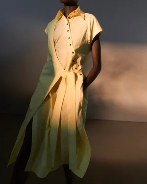 Model wearing a yellow button-up dress with a draped belt panel in a studio setting with soft lighting.