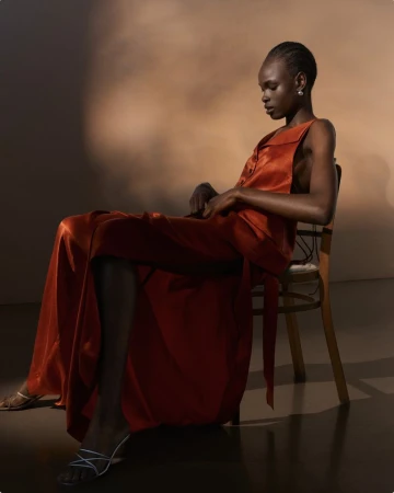 Model wearing a glossy orange satin dress seated on a wooden chair against a warm studio backdrop.