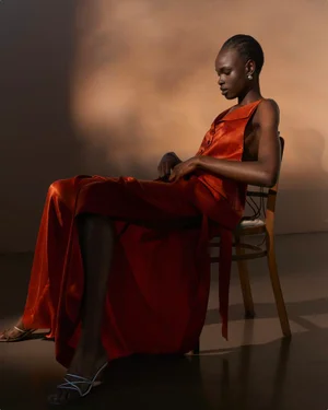 Model wearing a glossy orange satin dress seated on a wooden chair against a warm studio backdrop.