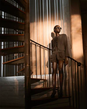 Model standing on a spiral staircase wearing a grey jacket and shorts with sunglasses against a slatted window background