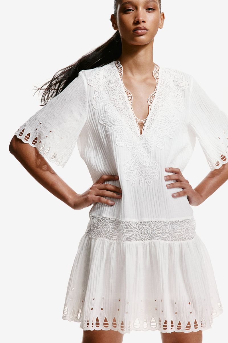 Model posing with hands on hips wearing a white embroidered lace-trimmed mini dress against a seamless white background.