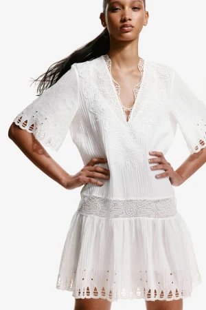 Model posing with hands on hips wearing a white embroidered lace-trimmed mini dress against a seamless white background.
